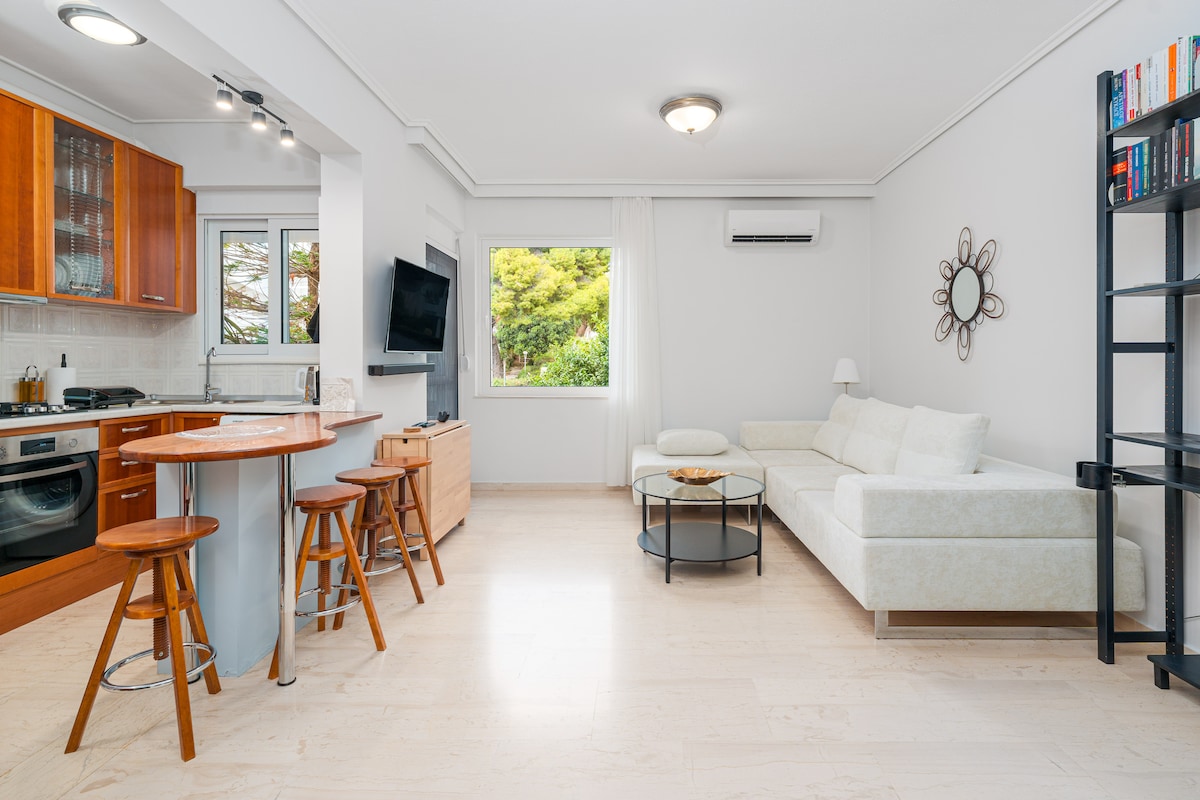 A bright living area features a spacious sectional sofa and a round coffee table. An open kitchen with wooden cabinets and bar seating is visible. Large windows allow natural light to fill the room, with greenery seen outside, providing a sense of tranquility.
