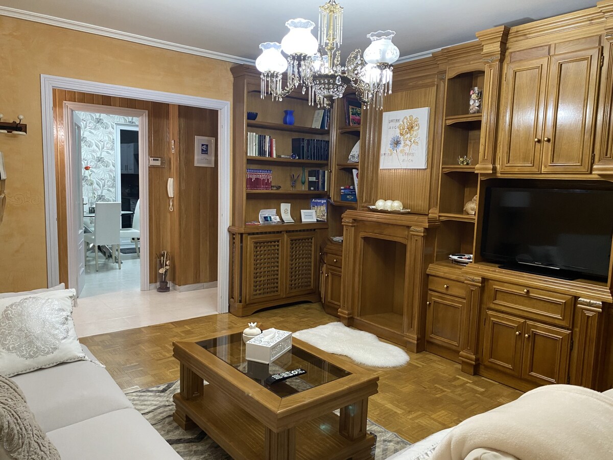 A living area is presented with warm wooden furnishings and a chandelier overhead. A coffee table occupies the center, accompanied by a plush rug. Shelves display books and decorative items, while a television is situated within a wooden cabinet. An arched entrance leads to a bright dining space.