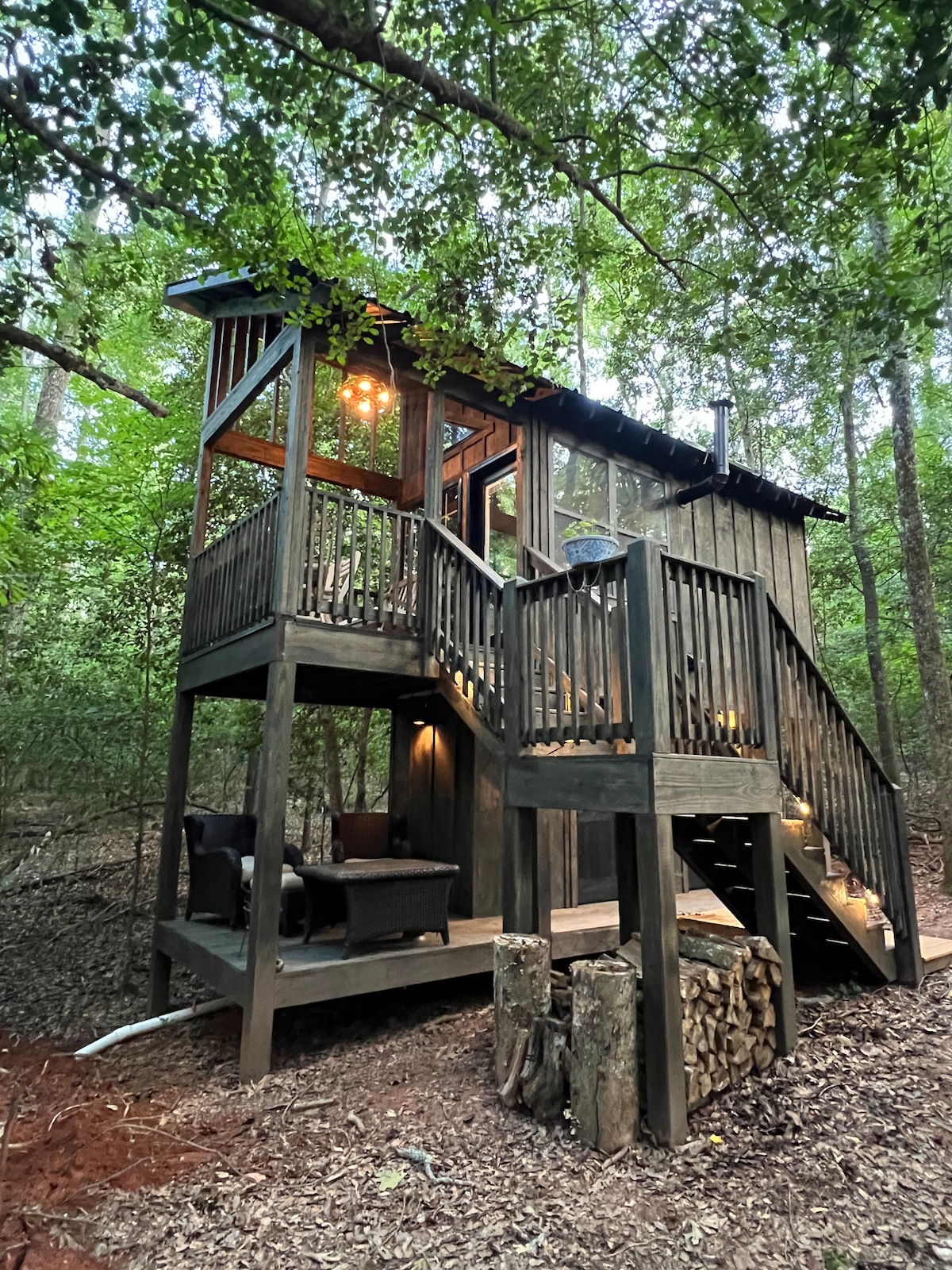 A modern treehouse is nestled among the trees, featuring a wooden exterior and a staircase leading to a deck. Comfortable outdoor seating is arranged on the porch. Lush greenery surrounds the structure, creating a serene woodland atmosphere.