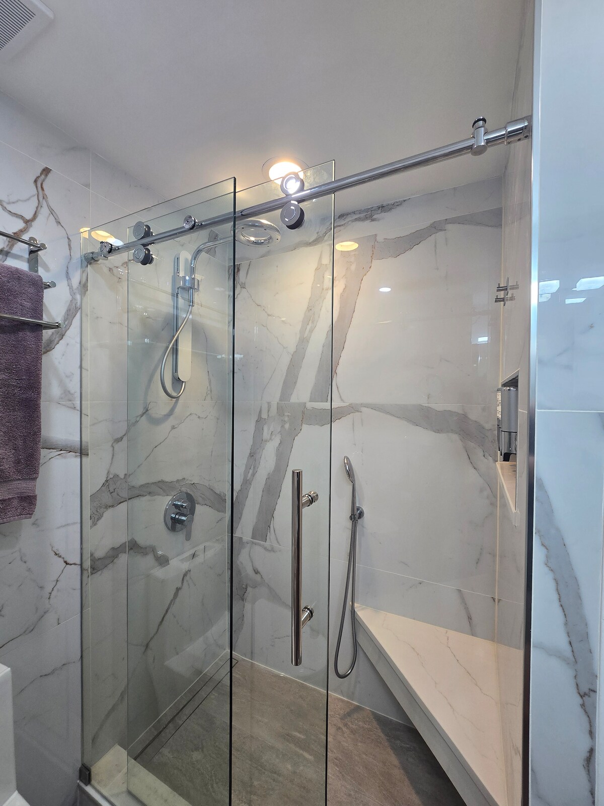 A modern spa shower features a glass enclosure with elegant marble walls. A rain showerhead is mounted overhead, while a handheld shower wand is also present. A built-in bench provides convenience, and neutral tones create a calming ambiance.