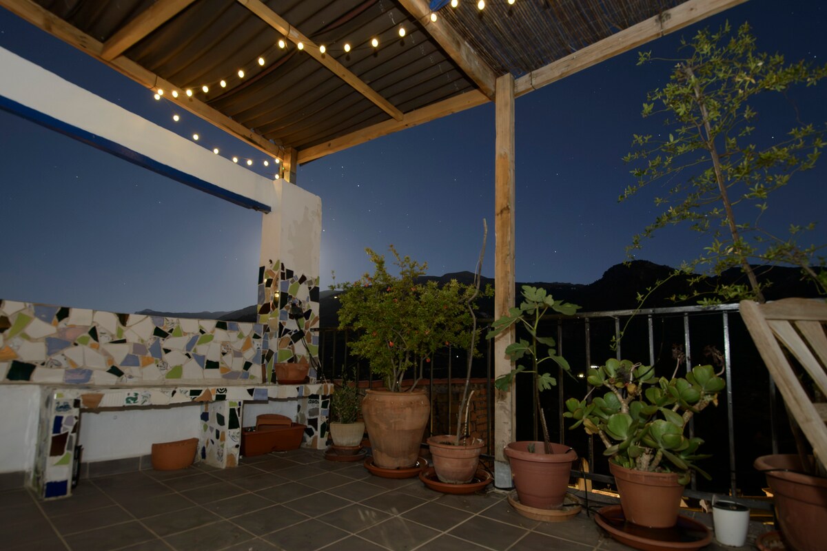 A spacious terrace is shown, featuring a handcrafted tile bench and various potted plants. Soft lighting from overhead string lights creates a cozy ambiance, while expansive views of the surrounding mountains and night sky add to the serene setting.