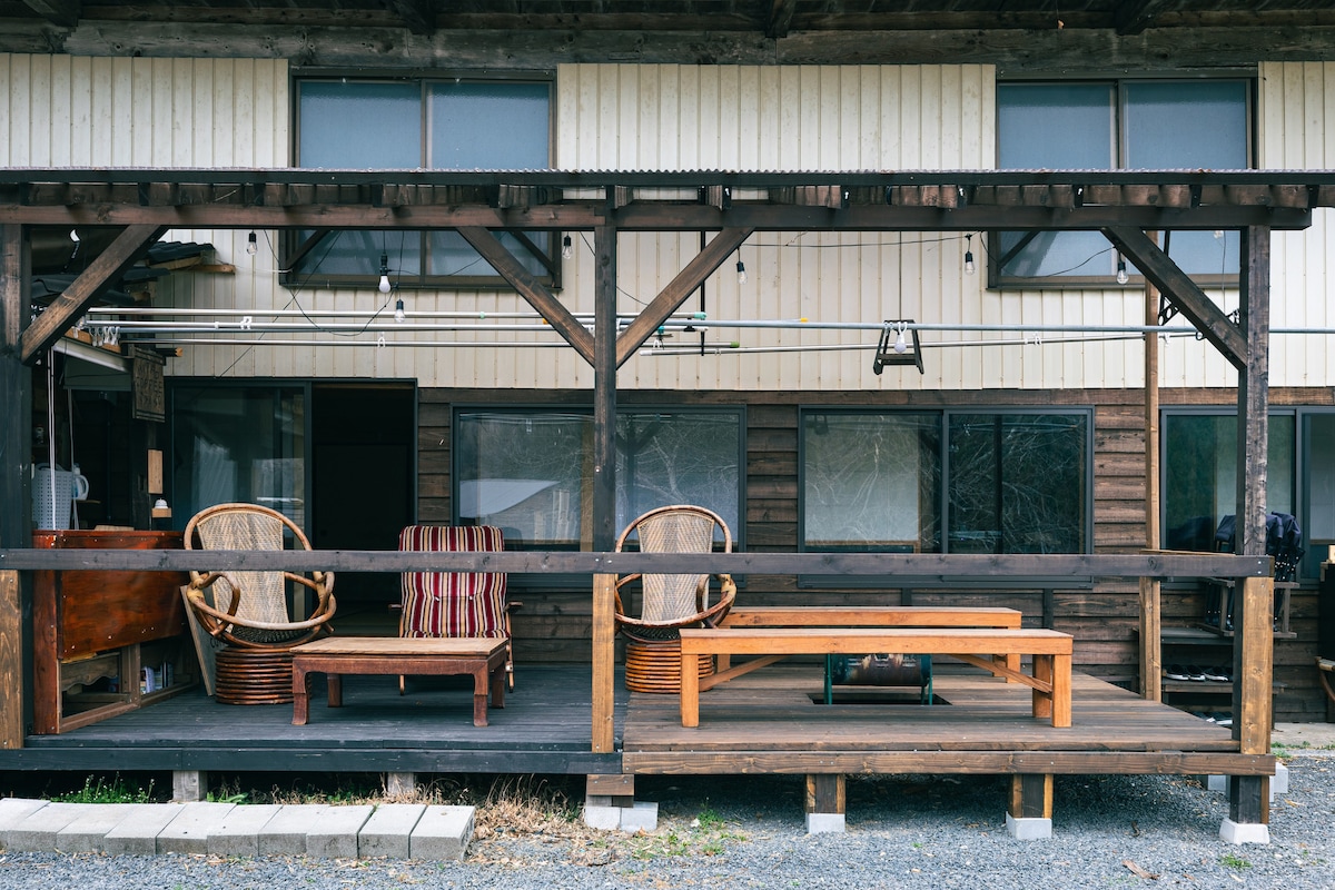 A covered porch area is presented with wooden beams and a wooden floor. Two rattan chairs and a coffee table are positioned for relaxation. Large windows behind the seating area offer views into the interior space.