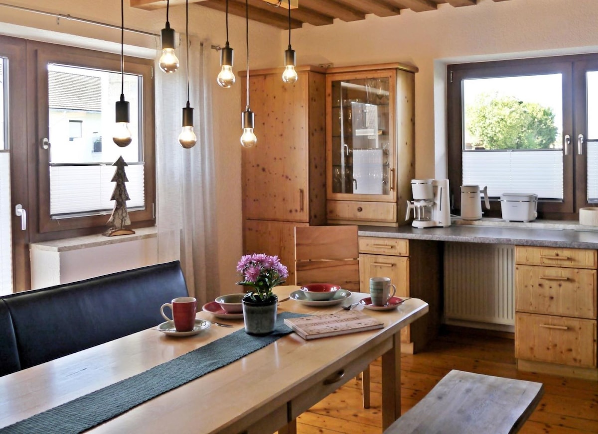 A wooden dining table is set with dishes and cups, accompanied by a small potted plant at the center. Warm light fixtures hang overhead, and natural light enters through two large windows. Wooden cabinets and a countertop are visible in the background.