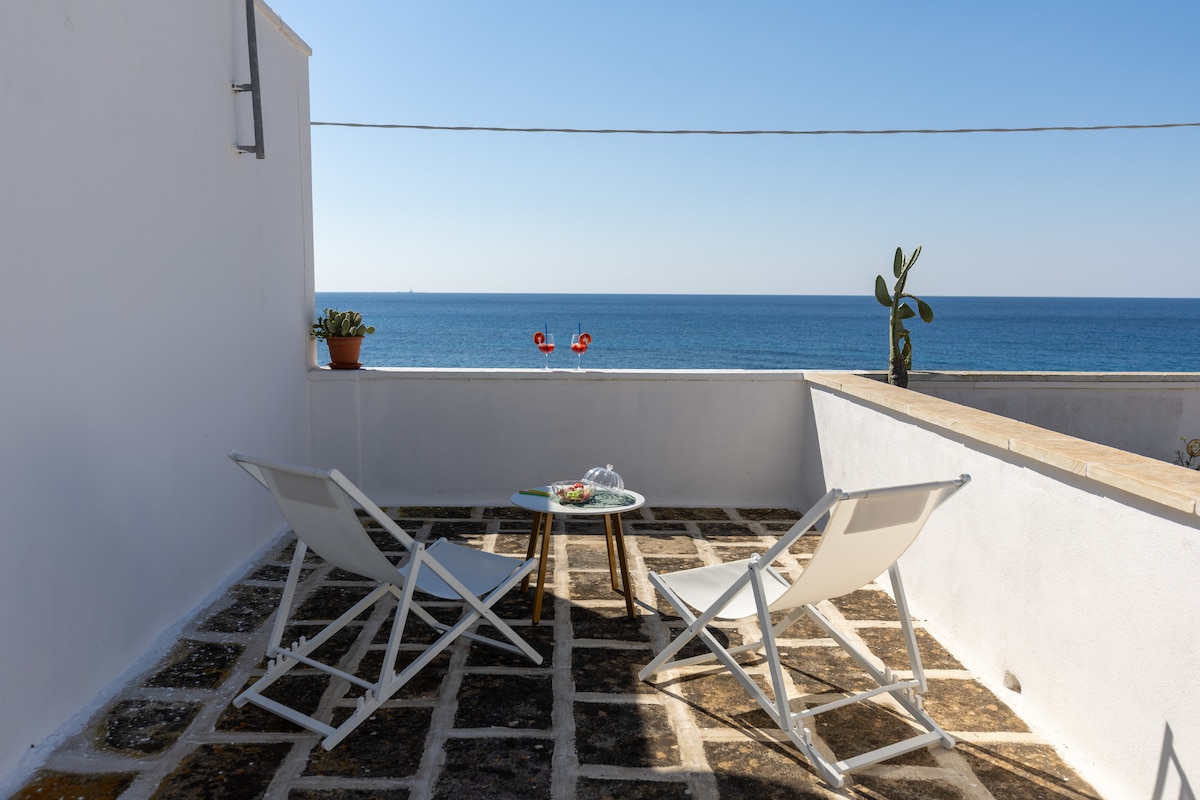 An outdoor terrace is presented, featuring a small round table and two white folding chairs. A potted plant adds a touch of greenery, while the serene sea is visible in the background under a clear blue sky.