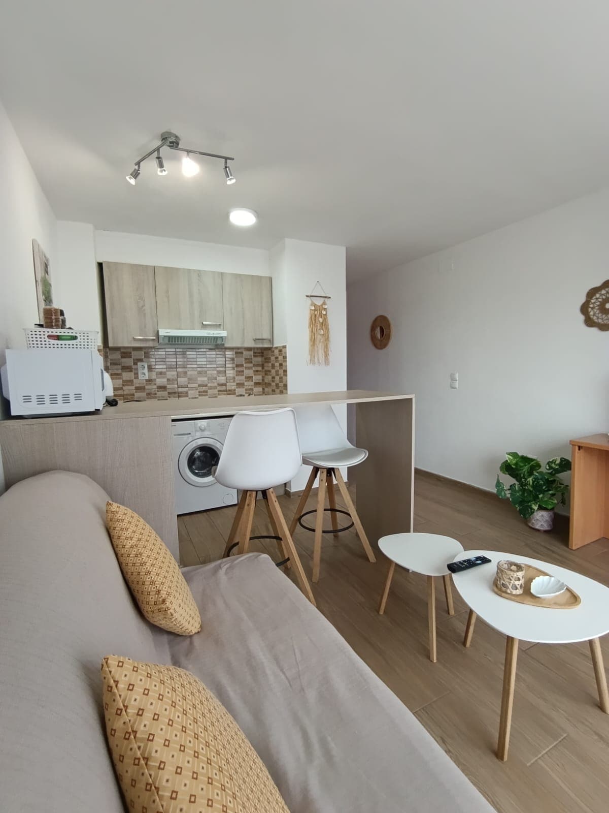 A compact living area features a light-colored sofa adorned with patterned cushions. A wooden table with two small stools is positioned nearby, with a kitchen area visible in the background. Modern appliances and a decorative plant are also present.