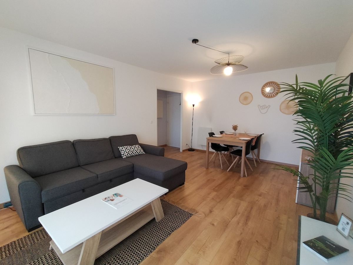 A spacious living area is presented, featuring a gray sectional sofa with patterned cushions. A light wooden coffee table is positioned in front, complementing the warm wooden flooring. A dining table for four sits nearby, accompanied by decorative wall accents and a floor lamp that provides ambient lighting.