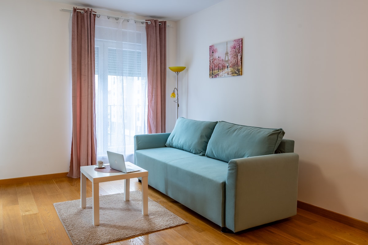 A cozy living area features a light blue sofa, positioned against a neutral wall. A small, white table is placed in front of the sofa with a laptop on it. Natural light filters through sheer curtains, and a piece of artwork is displayed above the sofa.