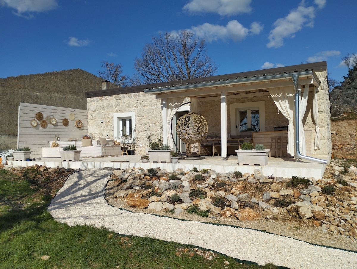 A charming stone house is set against a clear blue sky, featuring a spacious porch with comfortable seating and decorative elements. Lush greenery surrounds the property, while artistic wall decor adds character. The landscape includes a gravel pathway leading to the entrance.