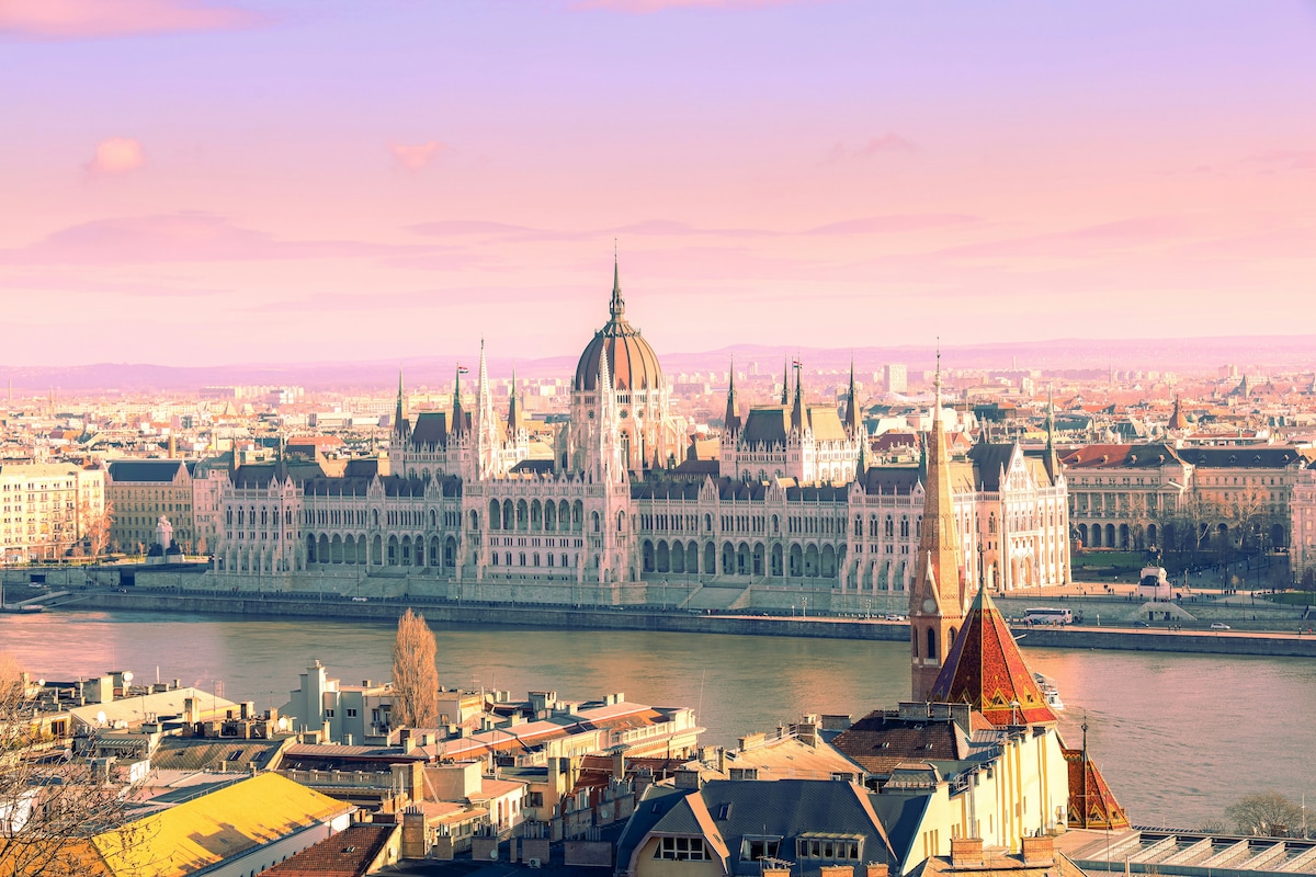 A panoramic view of the Hungarian Parliament Building is seen along the Danube River, displaying its impressive architectural features. The skyline is painted with soft hues of pink and purple as the sun sets, creating a serene backdrop for the vibrant cityscape below.