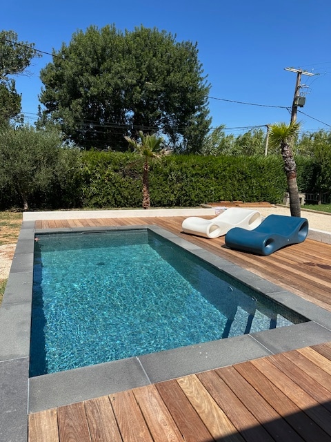 A private pool with clear blue water is framed by a wooden deck. Two lounge chairs in contrasting colors are positioned nearby, offering a place to relax. Lush greenery provides privacy around the pool area, contributing to a serene outdoor setting.