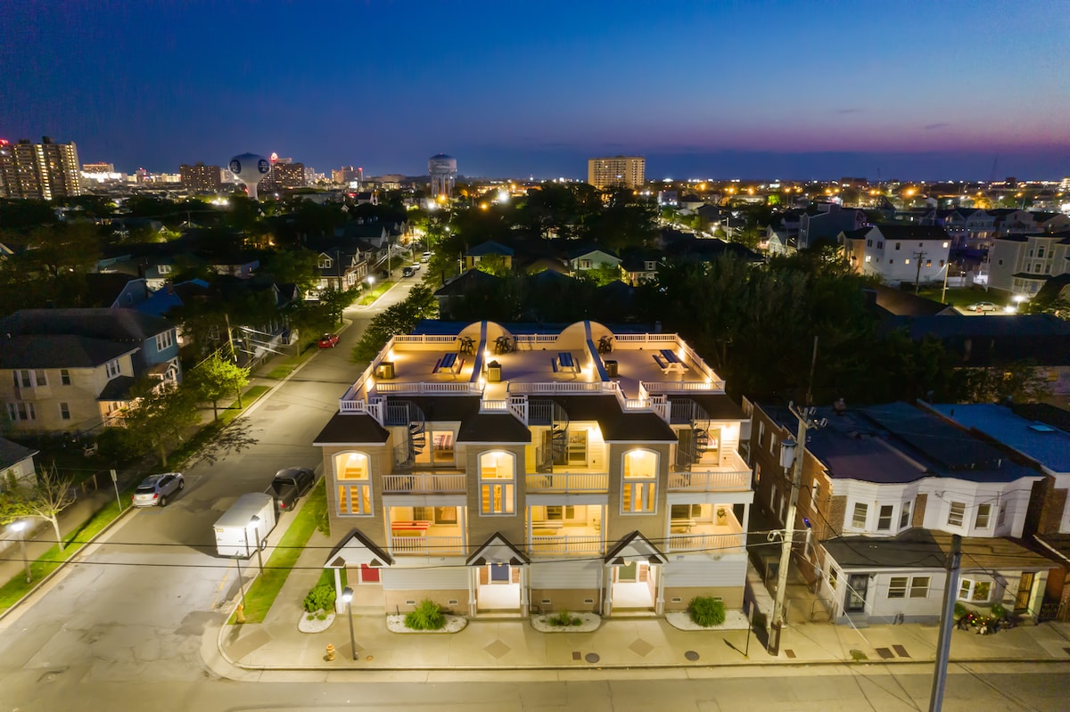 An aerial view captures the Top End Townhome at twilight, showcasing its modern architecture against a backdrop of a beautifully lit city skyline. The home features multiple rooftop decks, with warm lights illuminating the exterior, creating a welcoming atmosphere.