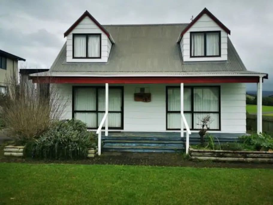A charming two-story holiday home is shown, featuring a symmetrical design with a light exterior and a sloped roof. Two large windows on the upper level are framed by dark accents. A welcoming porch area with steps leads to the front entrance, surrounded by neatly maintained greenery.