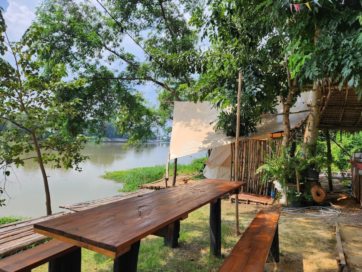 A wooden table with benches is set beside the Mae Klong River, surrounded by lush greenery and trees. A canvas shelter offers shade, creating a comfortable outdoor dining area that overlooks the calm waters and tranquil landscape.