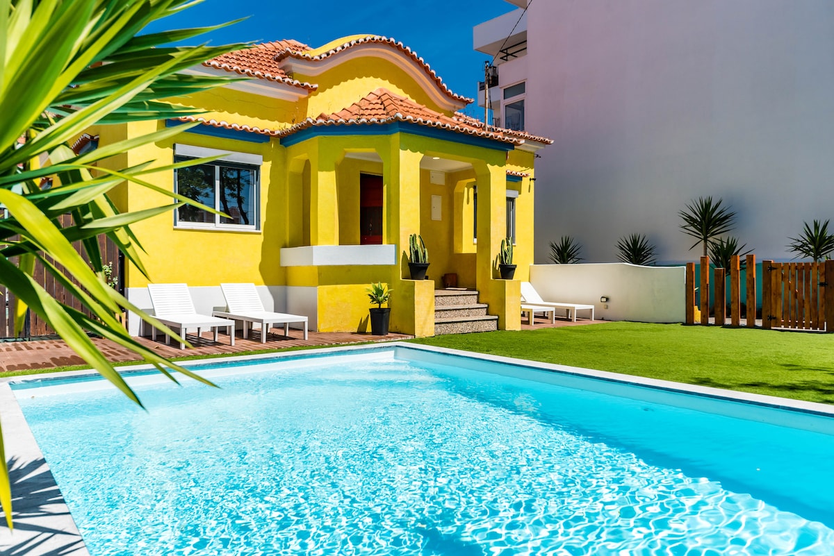 A vibrant yellow villa is framed by a clear, inviting swimming pool. Sun loungers are positioned near the poolside, and lush green grass surrounds the area. The architectural design features a tiled roof and large windows, creating an airy outdoor atmosphere.