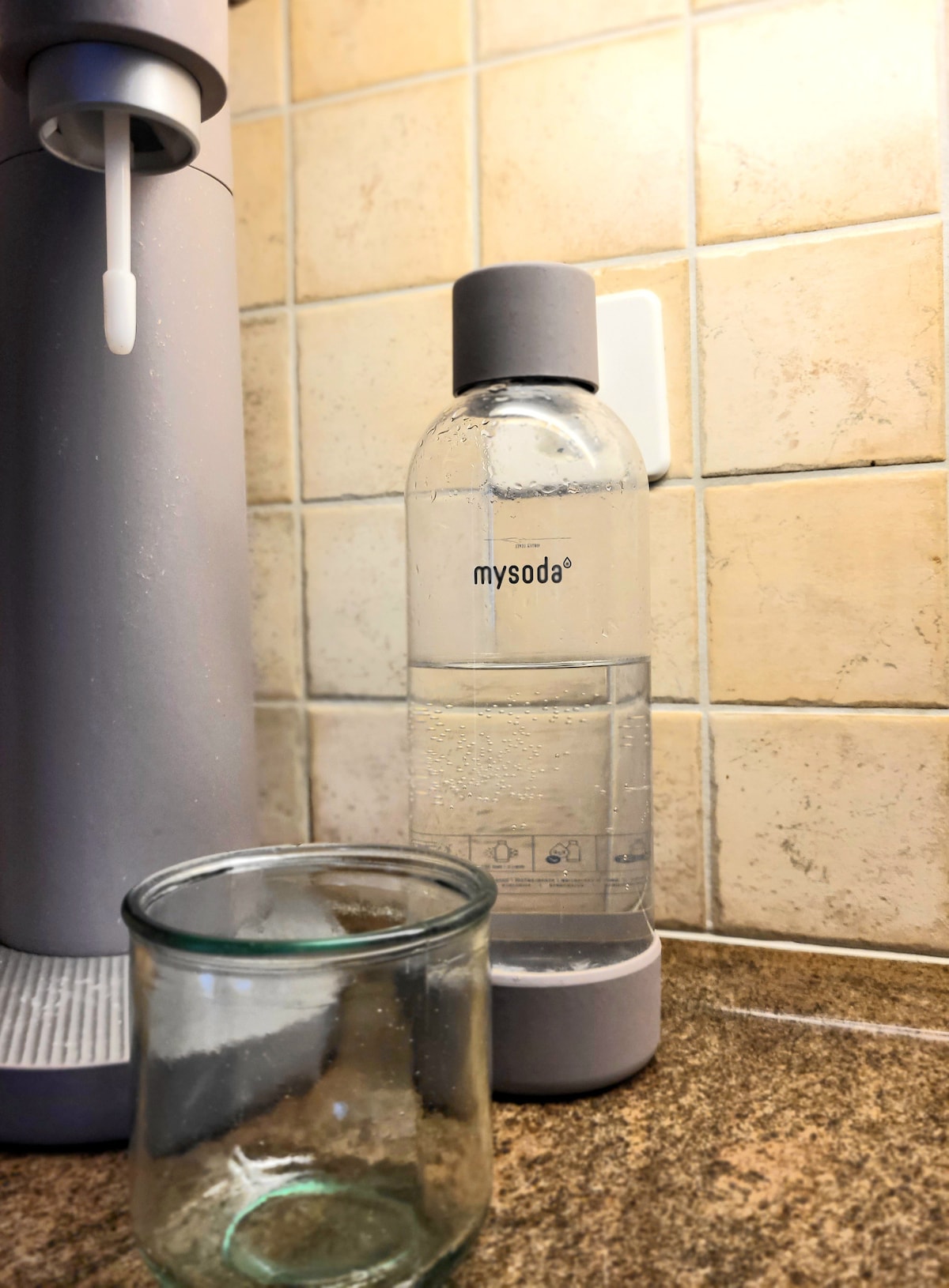 A soda maker is positioned beside a glass containing a small amount of water. The device is sleek and modern, featuring a clear plastic bottle with a gray cap. The background shows textured tiles, contributing to a clean and functional kitchen environment.