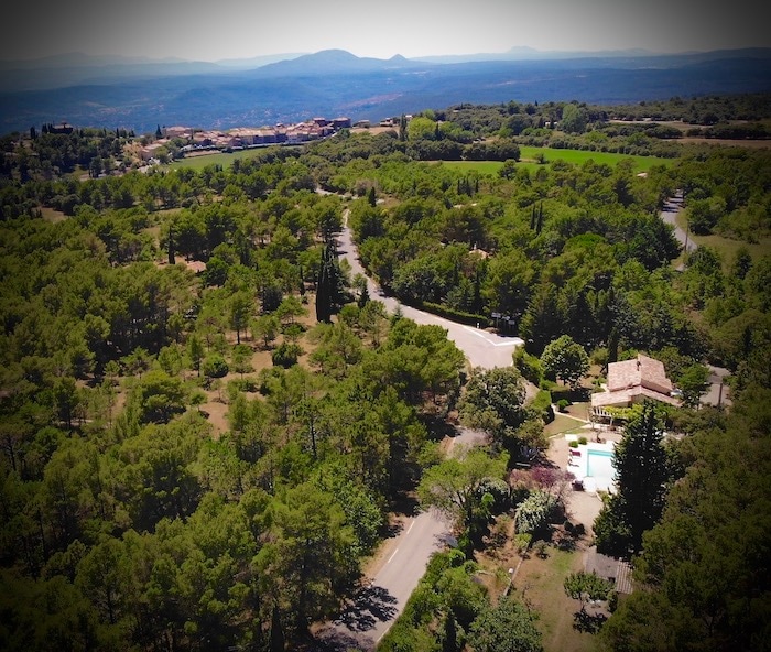 An aerial view captures the serene landscape surrounding the accommodation. Lush green trees and winding paths create a natural setting, with a glimpse of a swimming pool among the foliage. The village of Tourtour is faintly visible in the distance, enhancing the peaceful ambiance.