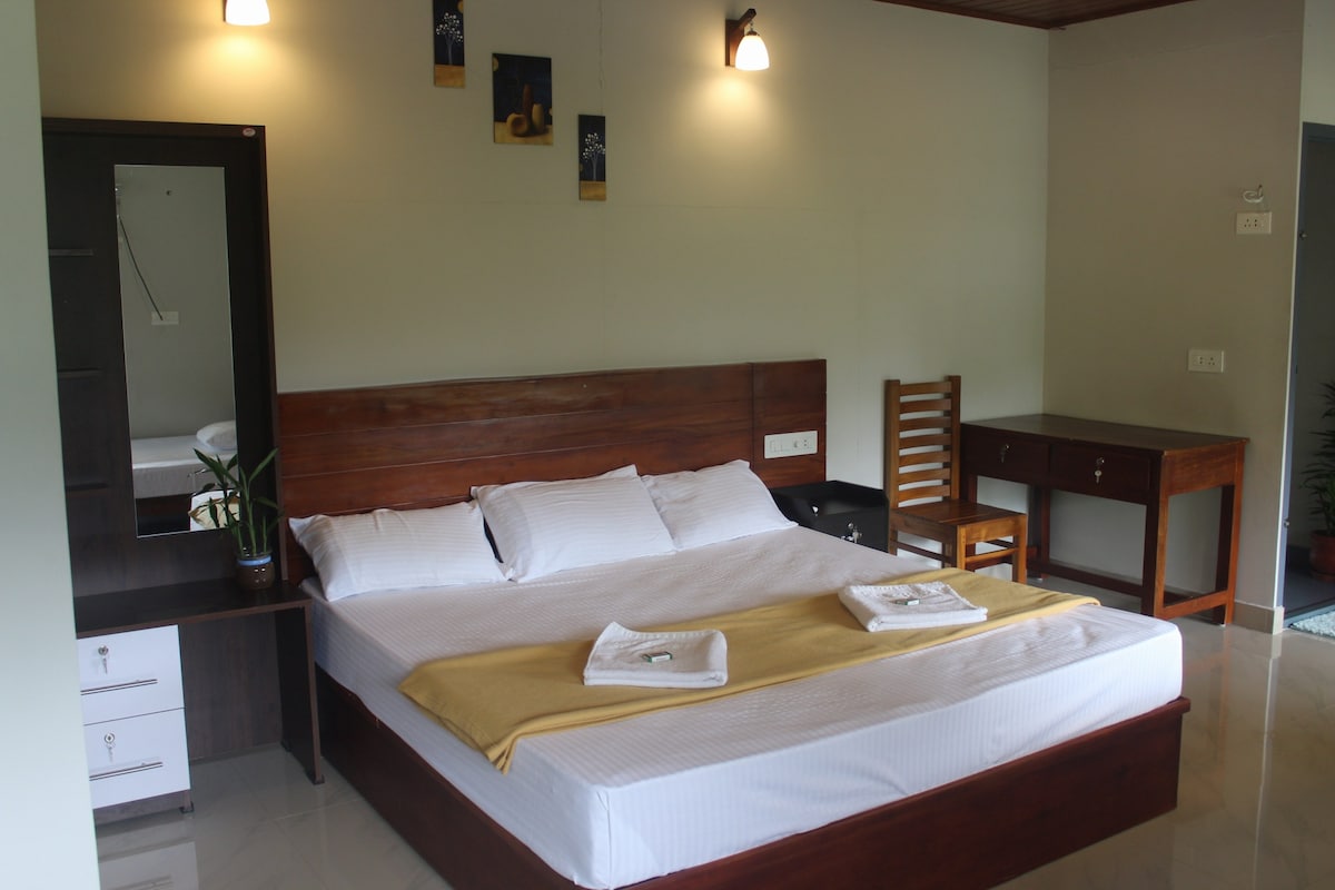 The sleeping area features a wooden bed with white linens and a yellow throw. A desk and chair are situated beside the bed, and a mirror is mounted on the wall opposite. Soft lighting is provided by wall sconces, creating a calm ambiance.