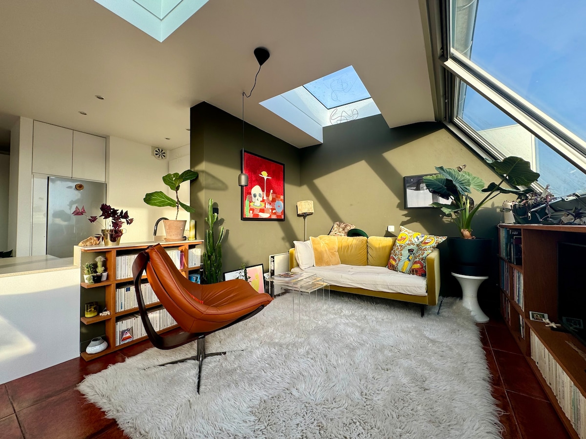 A spacious living area features a large rug, a modern chair, and a bright yellow sofa adorned with cushions. A skylight allows natural light to illuminate the room, complemented by decorative plants and art on the walls, creating a vibrant and welcoming space.