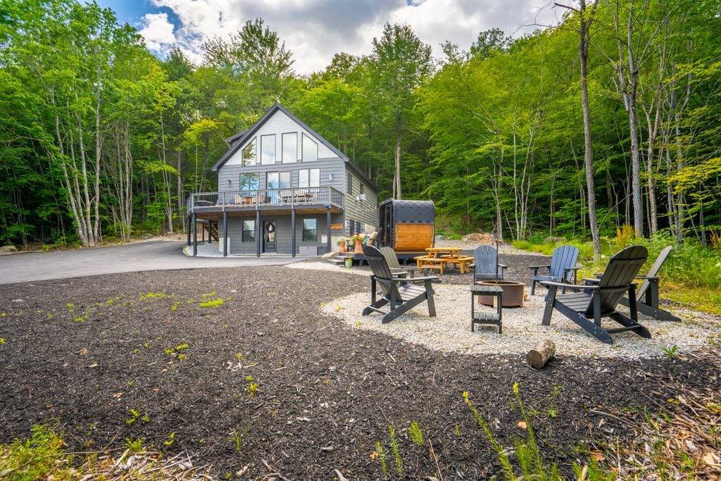 A modern home is surrounded by lush greenery, with an outdoor seating area featuring black adirondack chairs arranged around a fire pit. A custom sauna and seating area are visible, enhancing the spacious back yard, which provides access to the home’s elevated deck.