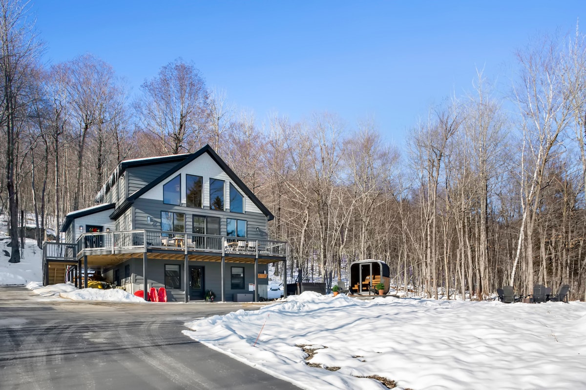 A modern home nestled among trees is visible, showcasing a large deck with outdoor seating. The snow-covered ground provides a serene contrast to the building's dark exterior. A storage shed is seen in the background, adding functionality to the winter landscape.