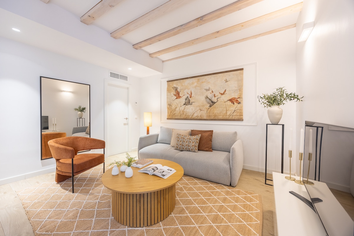A bright living area features a soft, light grey sofa paired with an orange accent chair. A round wooden coffee table sits centrally on a patterned rug, enhanced by a neutral artwork display above the couch. Soft lighting and wooden beams add warmth to the space.