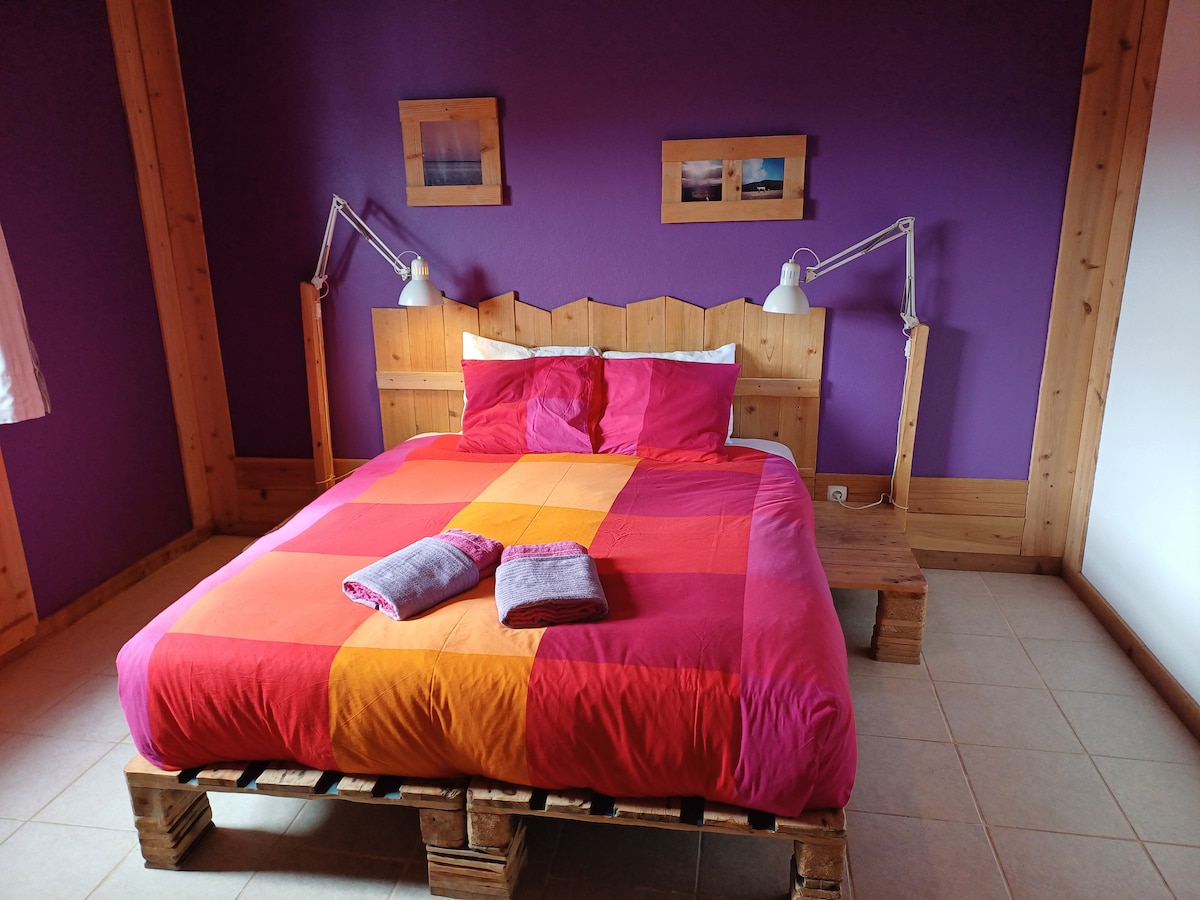A vibrant bedroom features a large bed with a patchwork duvet in shades of pink and orange. Two bedside lamps are mounted on the walls, and a wooden headboard adds a rustic touch. Light-colored tiles create contrast against the purple walls.