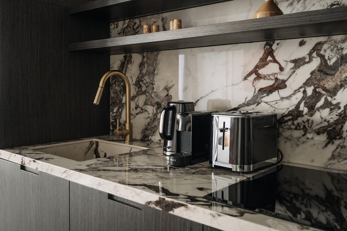 A modern kitchen countertop featuring a sleek marble surface, accentuated by a gold faucet. A kettle and a toaster are placed neatly beside each other against the backdrop of dark wood cabinetry and an artful marble wall.