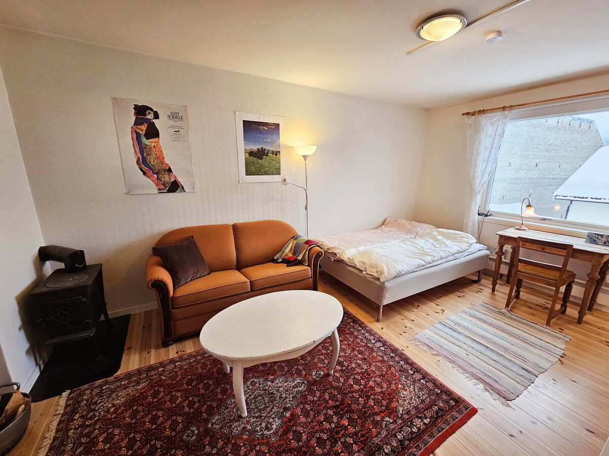 A combined living and sleeping area features a cozy sofa and a small white table. A spacious bed is positioned against the wall, covered with a light blanket. Decorative wall art and a window with sheer curtains allow natural light into the room.