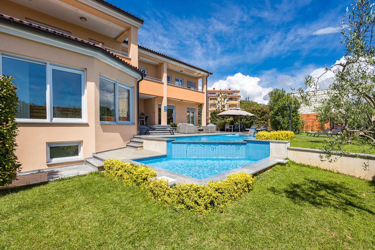 An inviting outdoor space features a clear blue pool surrounded by lush greenery and neatly trimmed hedges. The house’s light-colored exterior is complemented by expansive windows, providing plenty of natural light. A shaded seating area can be seen in the vicinity.