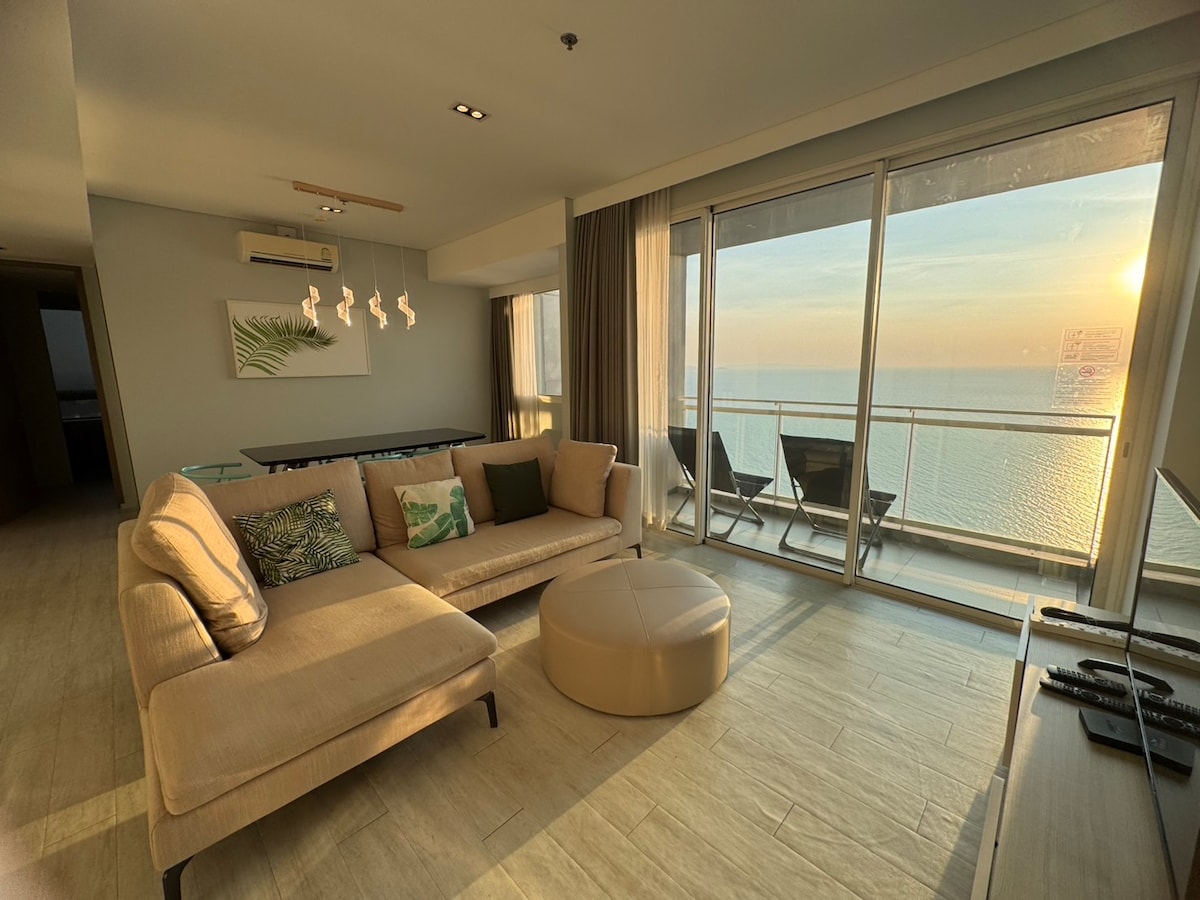 A spacious living area is highlighted by large glass doors offering views of the sea at sunset. A sectional sofa with assorted cushions faces a low ottoman. Natural light fills the room, complementing the modern furnishings and neutral color palette.