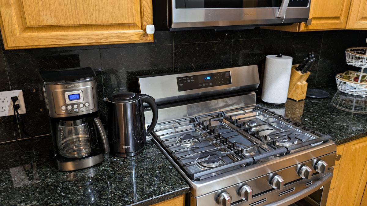 The kitchen features a sleek stainless steel gas stove, paired with a modern coffee maker and a kettle. Dark granite countertops provide an elegant backdrop, while a roll of paper towels and wooden utensils are neatly arranged for convenience.