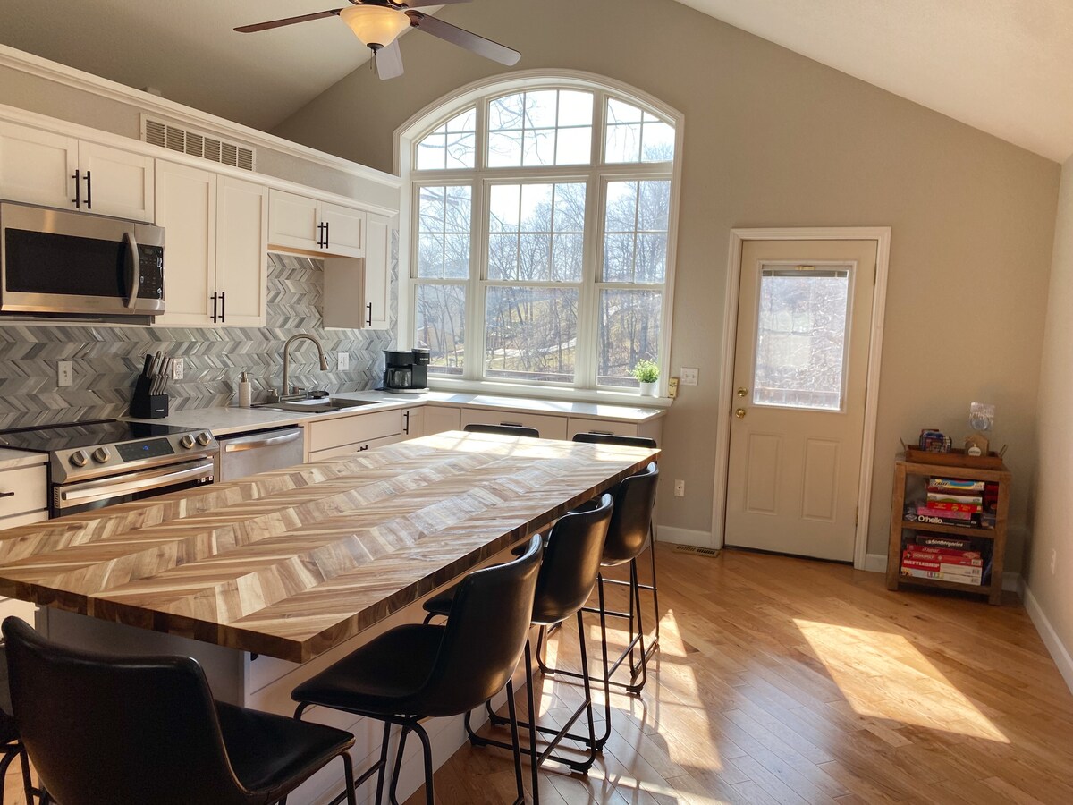 An open kitchen area features a large wooden island with seating for several guests. The white cabinetry is complemented by stylish appliances and a patterned backsplash. Natural light pours in through expansive windows, casting warm tones across the hardwood floor.