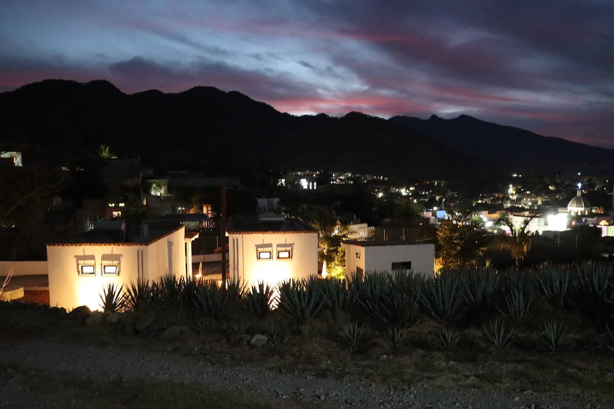 The image showcases a hillside view of illuminated white buildings against a backdrop of mountains at dusk. The sky features soft, colorful hues transitioning from daylight to night, with city lights twinkling below, creating a serene and picturesque atmosphere.