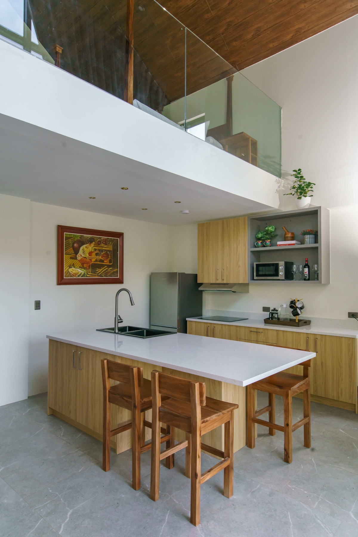 A modern kitchen features wooden cabinetry and a spacious island with seating for three. Stainless steel appliances are positioned against a wall, with a minimalist aesthetic complemented by a framed artwork above the island. Natural light enters from a window, enhancing the contemporary design.