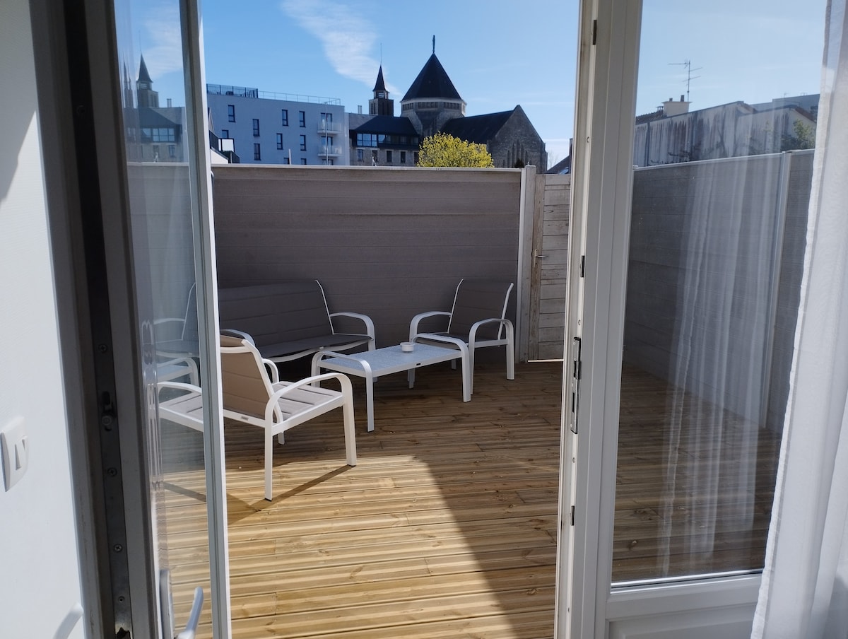 A private terrace is featured with wooden flooring and low-maintenance furniture, including two lounge chairs and a coffee table. A light color palette enhances the space, while the surrounding walls provide a sense of privacy. Sunlight streams in, suggesting a bright and airy environment.