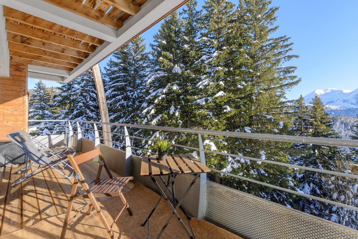 A spacious balcony is presented, featuring a table and two chairs, surrounded by tall pine trees blanketed in snow. The area offers a picturesque view of the mountain landscape, with sunlight illuminating the scene in a serene atmosphere.