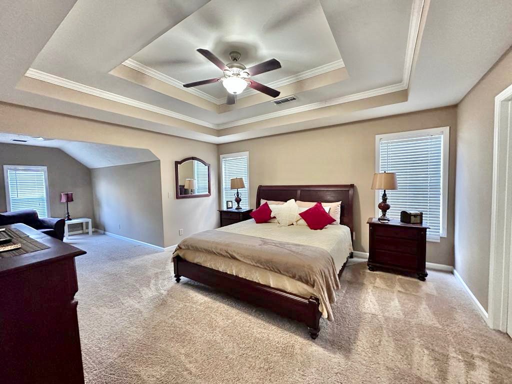A spacious bedroom is highlighted by a king bed with decorative pillows. Soft carpet covers the floor, and natural light filters through two windows. A ceiling fan provides air circulation. Nightstands with lamps and a dresser are positioned near the bed, enhancing functionality.
