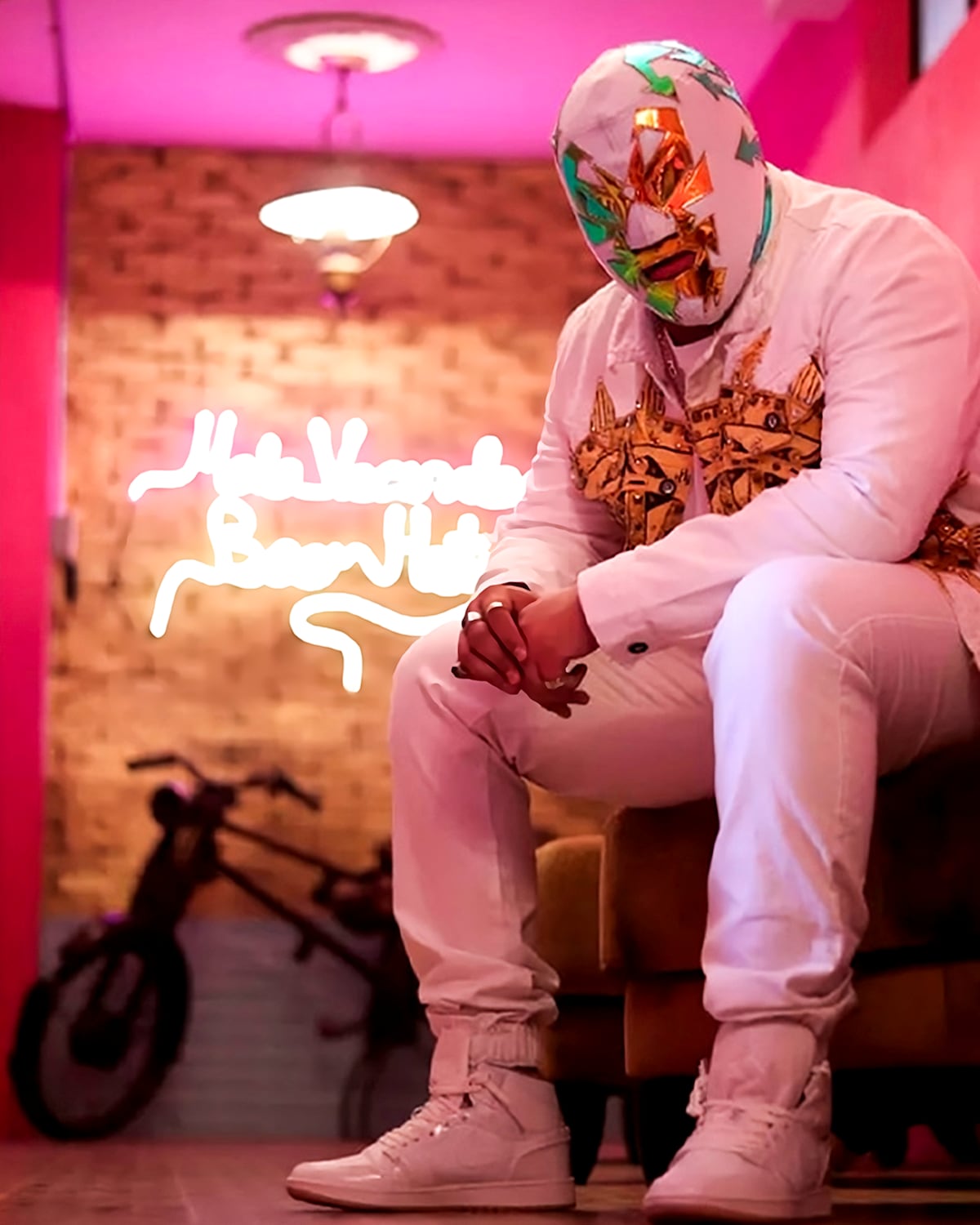 A seated individual in a vibrant room adorned with pink walls and artistic neon lighting. The figure wears a decorative mask and a white outfit, positioned beside a vintage bicycle that is partially visible in the background.