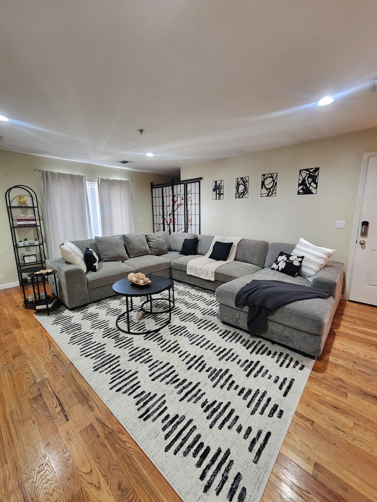 A spacious living area is presented, featuring a comfortable grey sectional sofa with decorative pillows. A round black coffee table is centered on a patterned area rug, while light-colored curtains frame large windows. Wall art and a decorative shelving unit enhance the warm ambiance.
