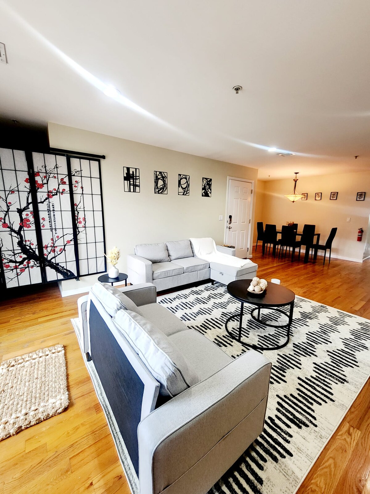 A spacious living area features modern furnishings, including a sofa and armchair arranged around a round coffee table on a patterned rug. Artful decor lines the wall, while a dining area with a table and chairs is visible in the background.