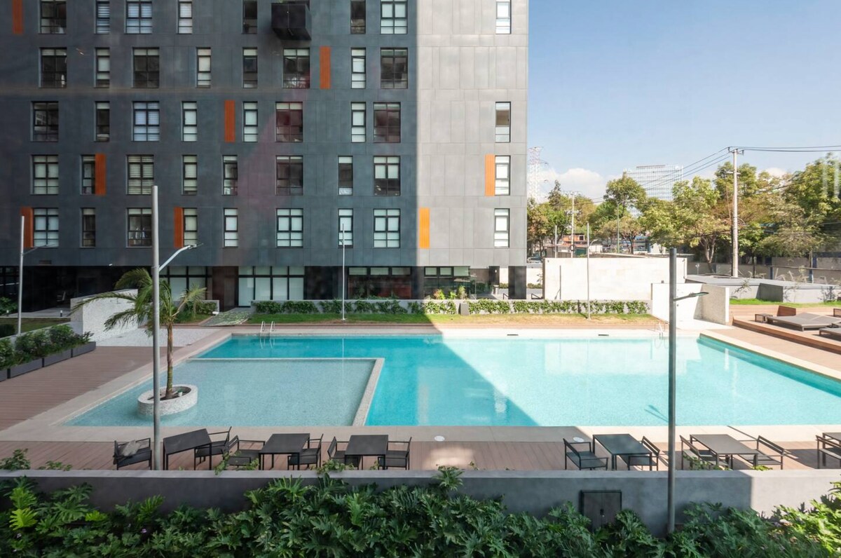 An outdoor swimming pool is surrounded by modern lounge chairs and palm trees. The sleek exterior of a multi-story building is visible in the background, complemented by vibrant orange accents. Clear blue water reflects the sky, creating a refreshing atmosphere.