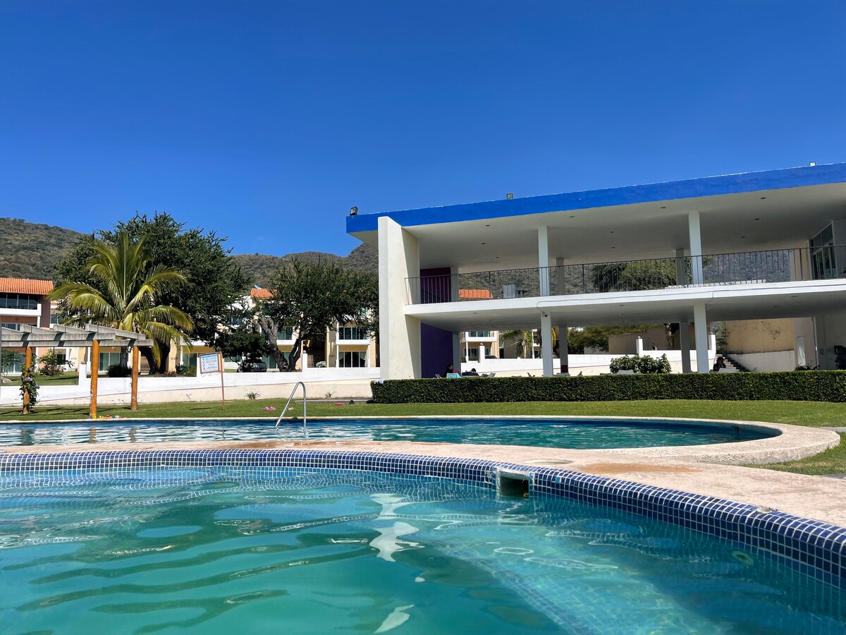 A clear swimming pool reflects the blue sky and is surrounded by well-manicured grass. A modern multi-story building stands in the background, featuring a white facade and a vibrant blue roof. Palm trees provide a touch of greenery near the pool area.