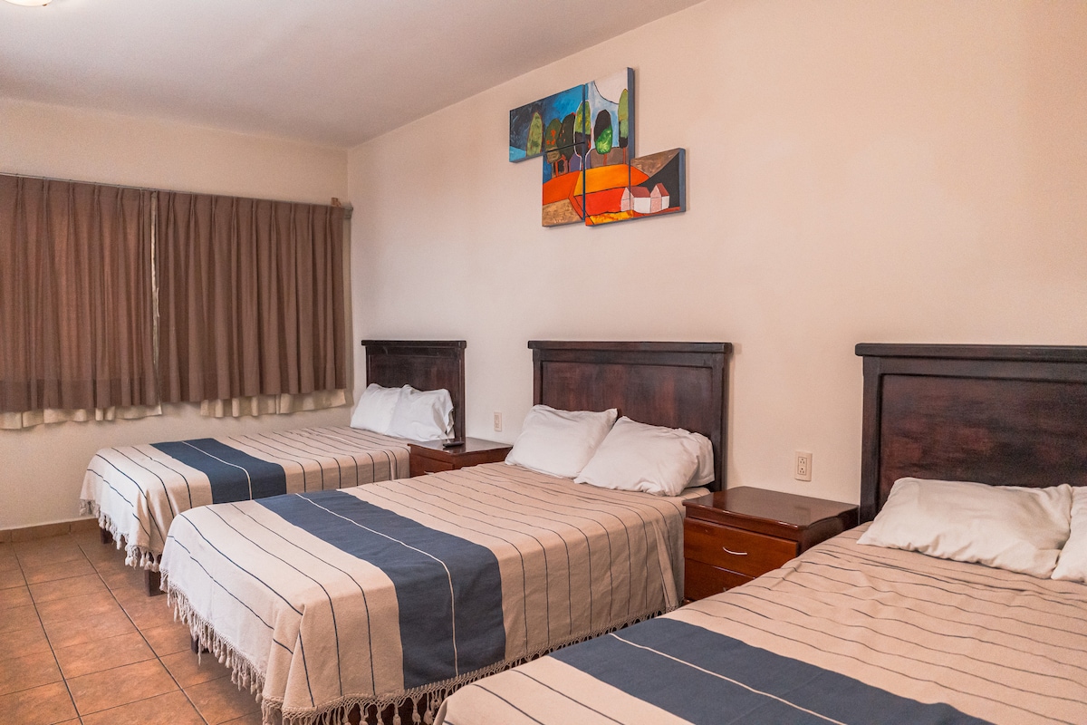 A spacious room features three beds with light-colored blankets. Dark wood nightstands are placed beside each bed. A colorful wall art piece is displayed above the beds. The room is illuminated by natural light filtering through a curtain-covered window.