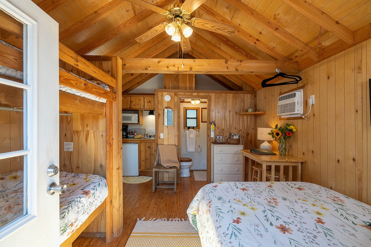 A wood-paneled interior showcases a cozy studio cabin with a full-size bed and bunk beds. A kitchenette is visible in the background, along with a private bathroom. Sunflowers adorn a small table, adding a bright touch to the space. Light filters through a nearby window.