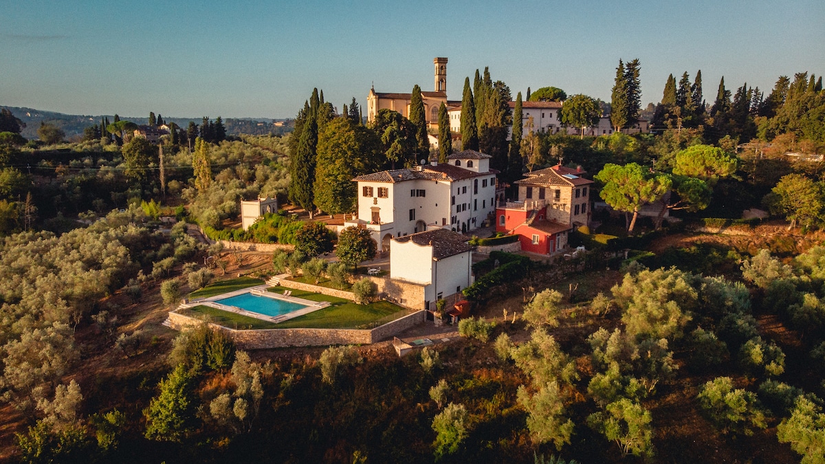 The image presents a charming rural village enveloped by greenery. A clear, inviting pool can be seen in the foreground, surrounded by manicured lawns. The buildings, displaying a blend of traditional architecture and lush landscaping, are set against a backdrop of rolling hills and trees.