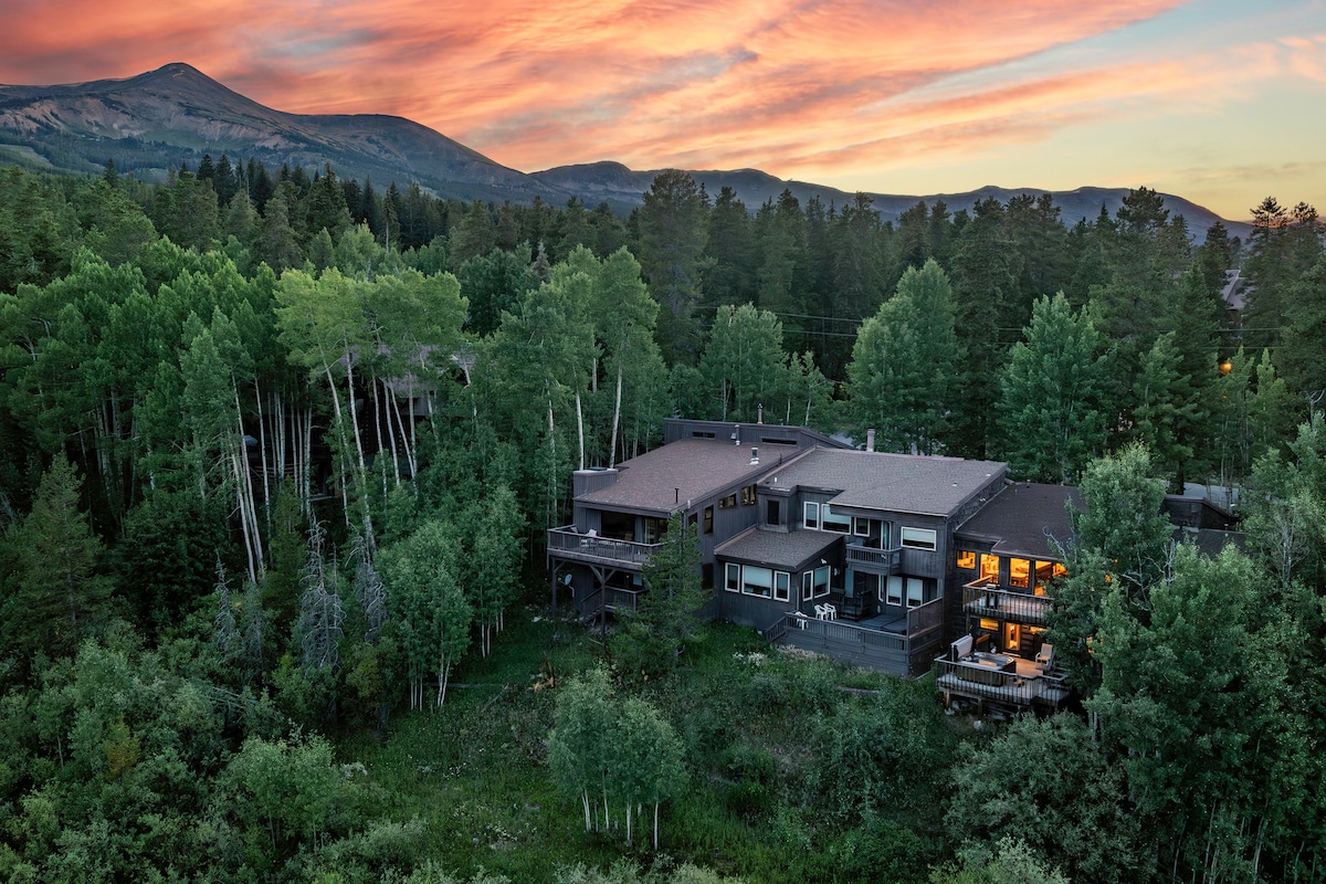The townhome is nestled amidst a lush forest, surrounded by tall trees, with a stunning mountain backdrop. A warm sunset sky casts a gentle glow over the property, highlighting its spacious structure and multiple decks that offer scenic outdoor views.
