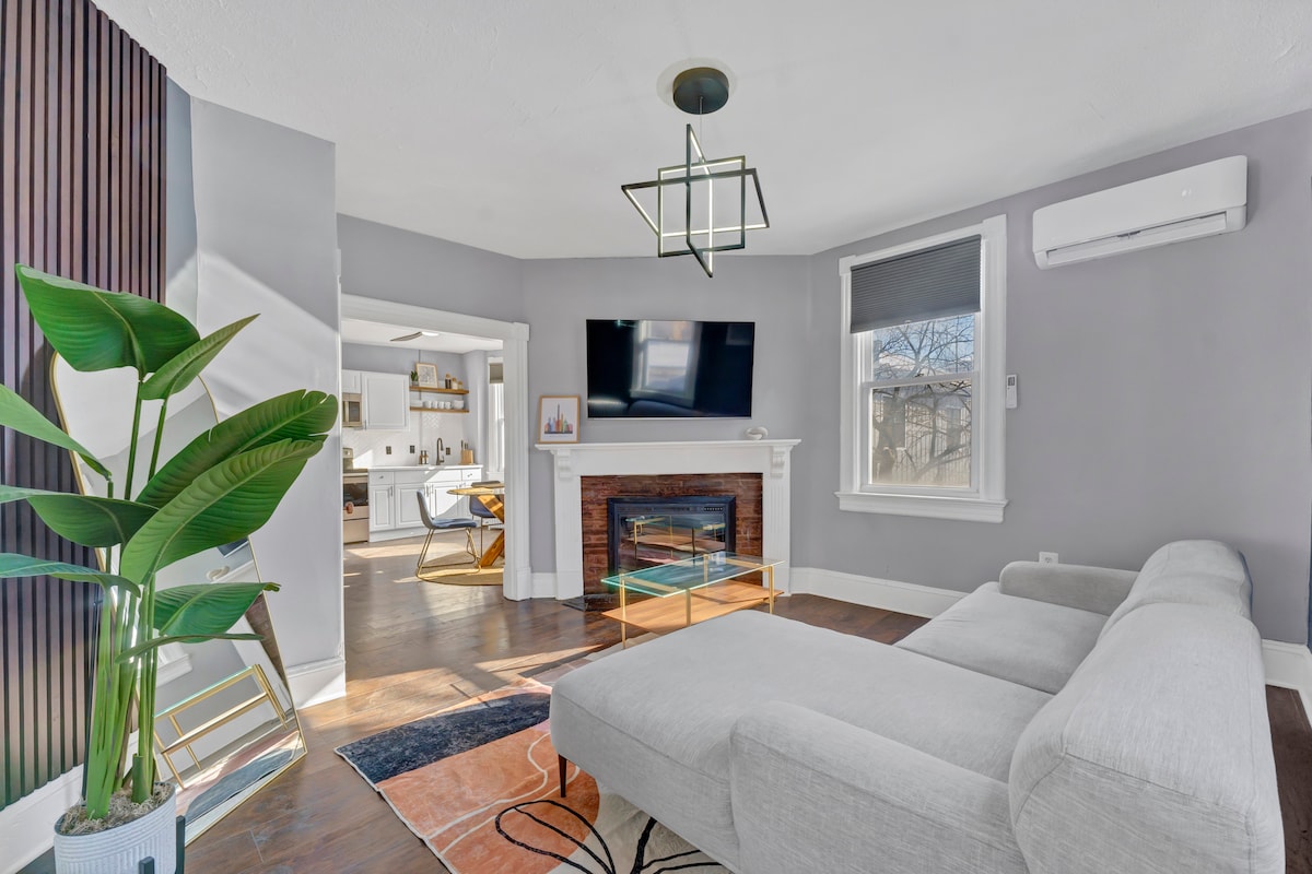 A stylish living area features a neutral sectional sofa facing a wall-mounted television. A modern light fixture hangs from the ceiling, and a decorative plant adds a touch of green. Light enters through a window with minimalistic coverings, creating a bright and open atmosphere.