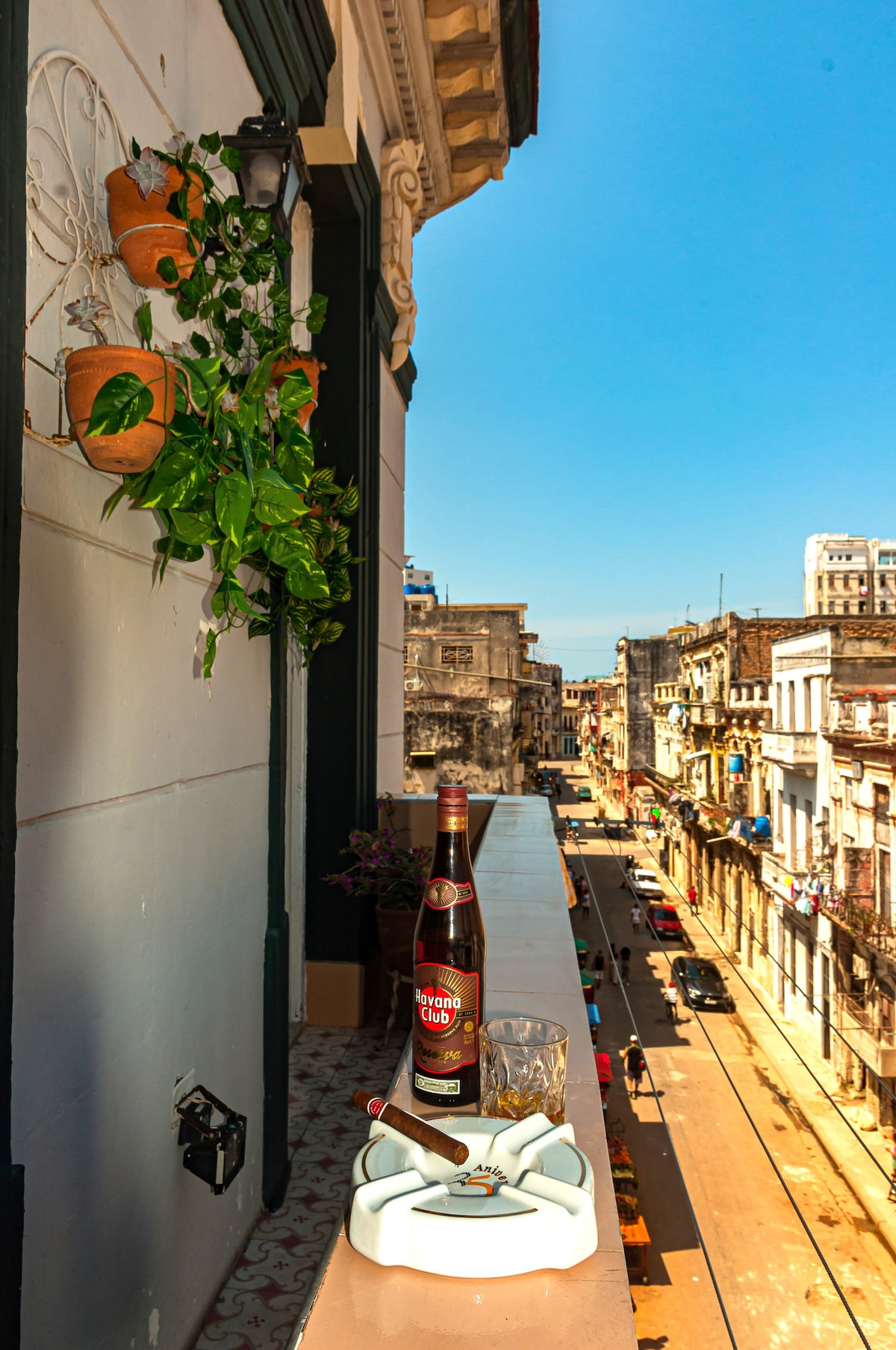 A sunlit balcony features potted plants alongside a tasteful wooden railing. A bottle and glass sit next to an ashtray, overlooking a vibrant street lined with historic buildings, capturing the essence of the lively atmosphere of La Habana.