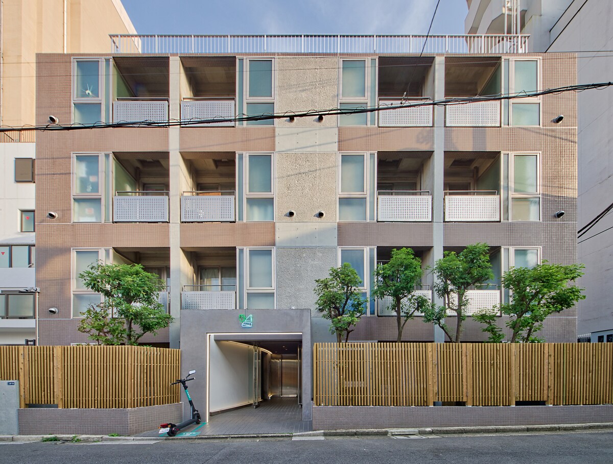 A modern apartment building is shown with a clean facade featuring large windows and balconies. Lush greenery is visible at the entrance, complemented by a wooden fence. A small pathway leads to the entrance, while a bicycle is parked nearby, enhancing the urban residential atmosphere.