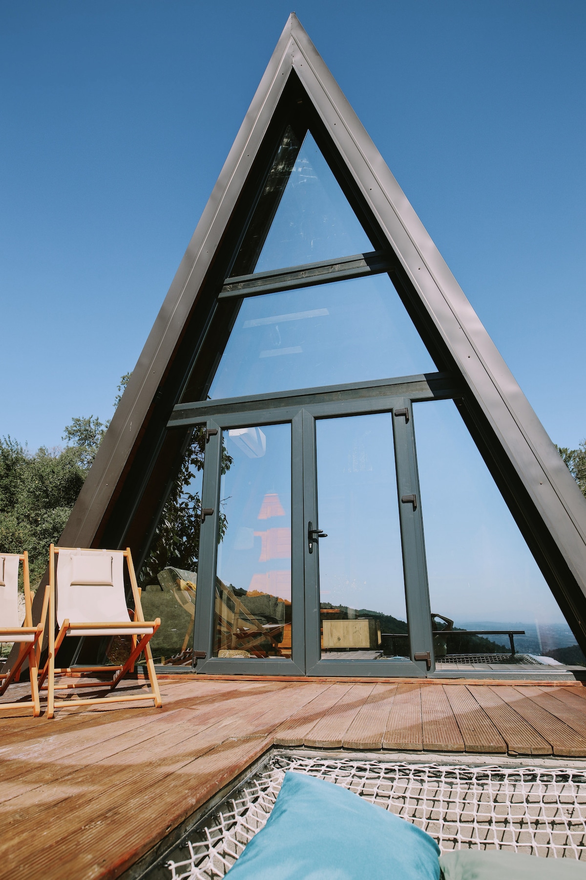 An A-frame cabin is showcased with large glass doors that create an open and inviting entrance. A wooden deck features two light-colored chairs for outdoor seating. The backdrop includes clear blue skies and glimpses of nature, enhancing the cabin's serene setting.